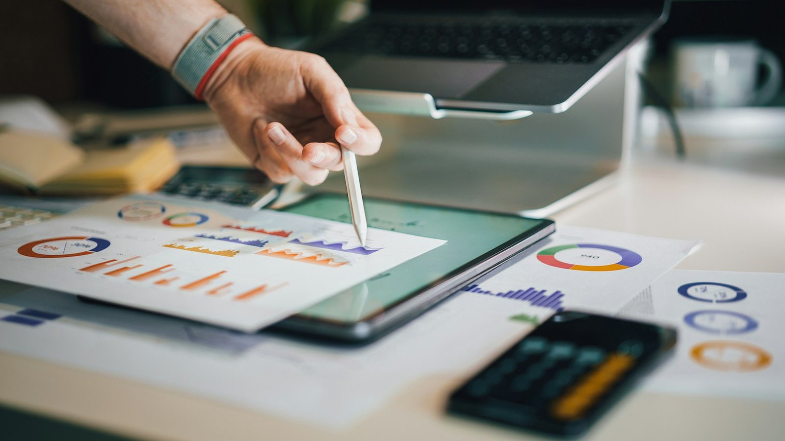 A hand reviewing marketing charts on a tablet beside printed reports and a calculator